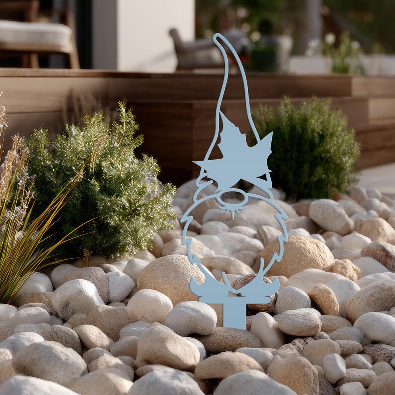 May include: A light blue metal garden gnome silhouette. The gnome has a long pointed hat with a maple leaf design, a beard, and a round nose. It is placed in a bed of rocks and pebbles, with greenery in the background. The gnome is approximately 12 inches tall.
