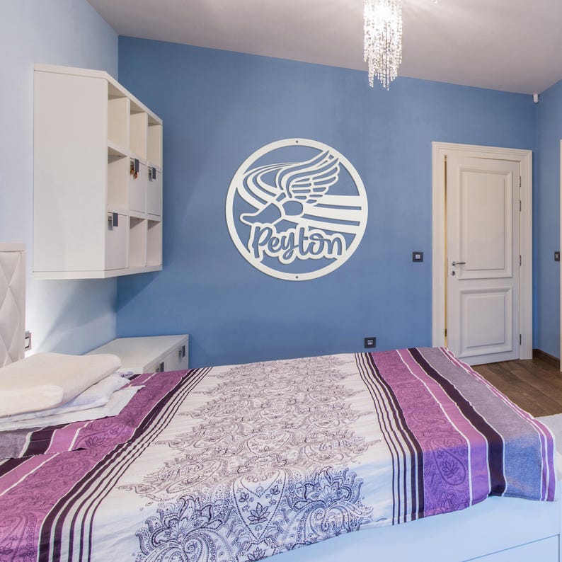 Puede incluir: Un letrero de pared circular blanco con un gr&aacute;fico de un pie alado y el nombre Peyton en letra cursiva, montado en una pared azul sobre una cama con un edred&oacute;n de estampado cachemir morado y blanco y una unidad de almacenamiento blanca.