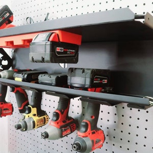 May include: A black metal wall-mounted storage rack with a fold-down shelf. The rack has several drill drivers hanging from it and two battery packs on the shelf. The battery packs are red and black and have the text "M18 FUEL LITHIUM-ION XC 4.0"