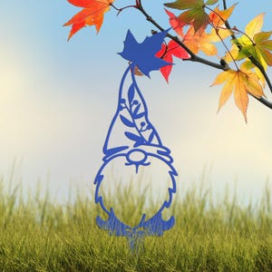 May include: A blue metal garden gnome stake with a beard and hat, topped with a blue leaf. The gnome is set in a grassy area with colorful autumn leaves in the background.