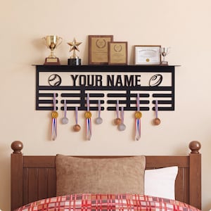 Puede incluir: Un expositor de pared de metal negro con el texto YOUR NAME, un icono de béisbol y un icono de balón de rugby. El soporte incluye varias barras horizontales para colgar medallas, con varias medallas de oro y plata suspendidas de cintas.