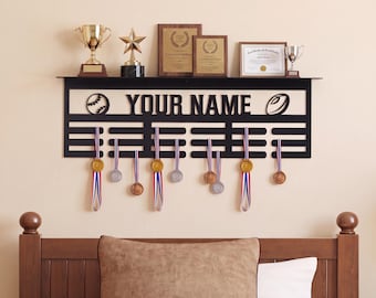 May include: A black metal wall-mounted display rack featuring the text YOUR NAME, a baseball icon, and a rugby ball icon. The rack includes multiple horizontal bars for hanging medals, with several gold and silver medals suspended from ribbons.