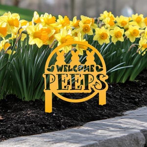 May include: A yellow circular garden stake featuring three bunny silhouettes at the top, two small hearts on the sides, and the text "WELCOME PEEPS" in the center. The stake is positioned in dark soil in front of a patch of bright yellow daffodils.