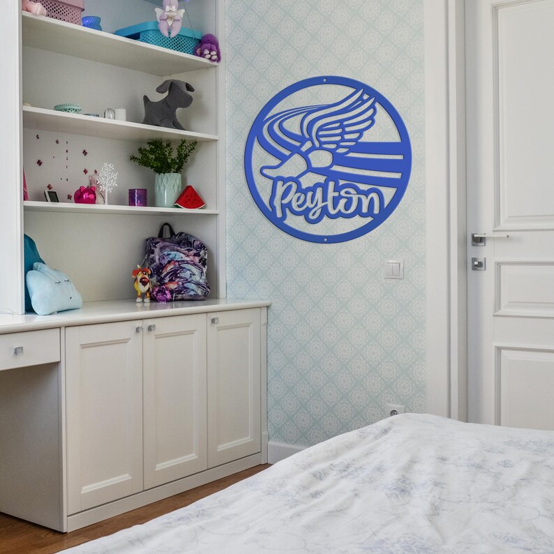 Puede incluir: Un letrero de pared circular azul que presenta un gr&aacute;fico estilizado de un zapato alado y el nombre Peyton en letra cursiva. El letrero est&aacute; montado en una pared de color claro con un patr&oacute;n geom&eacute;trico sutil, sobre un mueble blanco.