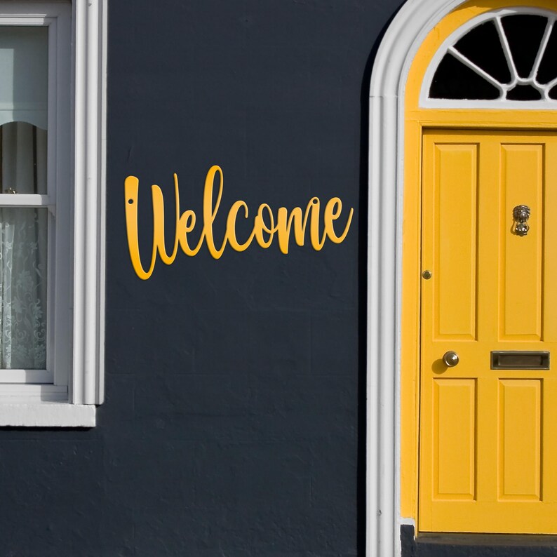 May include: A yellow "Welcome" sign is mounted on a dark blue exterior wall. A yellow door with a white arched frame is to the right. A window with white trim is on the left. The sign is in a cursive font.