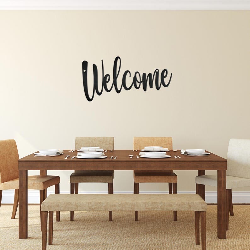 May include: A black metal "Welcome" sign is mounted on a beige wall above a dining table. The table is made of dark wood and is set with white plates and silverware. Beige chairs and a bench surround the table.