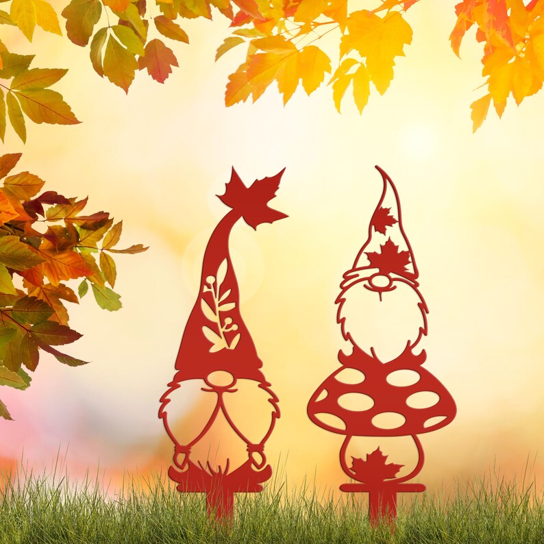 May include: Two red metal garden gnomes with autumn leaf designs. One gnome has a tall hat with a leaf, the other has a mushroom-shaped body and a hat with maple leaves. They are placed in a grassy area with fall foliage in the background.
