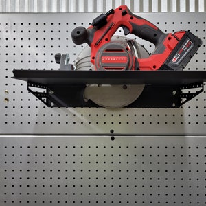 May include: A red and black circular saw with a black metal shelf mounted on a silver pegboard wall. The saw has a black handle and a black and silver blade. The saw is labelled "Brushless" and "18V MAX Lithium-Ion XC".