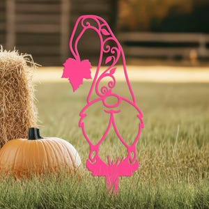 May include: A bright pink metal garden gnome stake with a leaf and swirl design. The gnome is standing in a grassy area with a pumpkin and hay bale in the background. The gnome is a decorative garden accent.