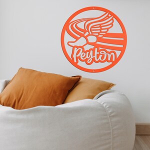 Puede incluir: Un letrero de pared circular de color naranja con un dise&ntilde;o recortado de un zapato alado con l&iacute;neas de movimiento. El nombre Peyton est&aacute; escrito en letras cursivas blancas en el centro del marco circular. El letrero est&aacute; montado en la pared.