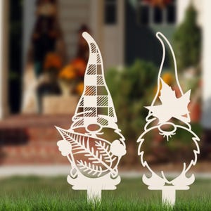 May include: Two cream-colored gnome yard stakes. One gnome has a plaid hat and holds a leaf. The other gnome has a leaf on its hat and a beard. Both have stakes for easy placement in the yard. Fall garden decor.