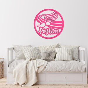 Puede incluir: Un letrero de pared circular de color rosa intenso que presenta un gr&aacute;fico estilizado de una zapatilla de correr alada y el nombre Peyton escrito en letra cursiva. El letrero tiene un dise&ntilde;o recortado y est&aacute; montado en una pared blanca.