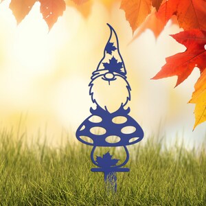 May include: A blue metal garden gnome stake. The gnome has a pointed hat with leaf cutouts, a beard, and a mushroom-shaped body with circular cutouts. The stake is designed to be placed in a garden or yard. The background features grass and autumn leaves.