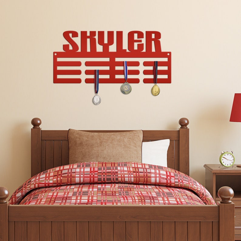 May include: Red metal medal display rack with the name "Skyler" in large letters. The rack has 10 slots for medals and three medals are hanging on the rack. The rack is mounted on a wall above a wooden bed frame with a red and white checked comforter.