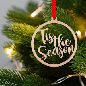 Könnte beinhalten: Ein hölzerner Weihnachtsschmuck mit dem Schriftzug "Tis the Season" in Schreibschrift. Der runde Schmuck hängt an einem roten Band. Er befindet sich auf einem grünen Weihnachtsbaum mit Lichtern.
