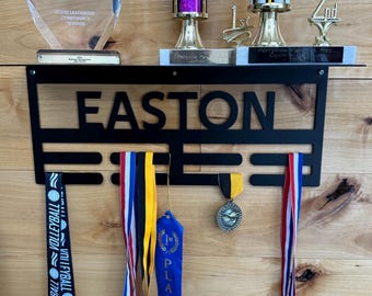 May include: A black metal medal display rack with the name Easton on it. The rack has three horizontal bars for hanging medals. There are four medals hanging on the rack, including a blue ribbon with the words "1st Place" on it, a black and white ribbon with the words "Volleyball" on it, and a yellow and black ribbon. There are also two trophies on a shelf above the rack.