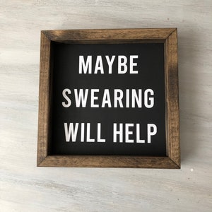 May include: A framed sign with a black background and white text that reads "MAYBE SWEARING WILL HELP". The frame is made of dark wood.