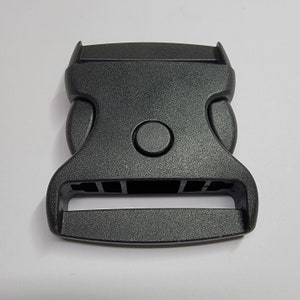 May include: A black plastic buckle with a smooth, textured surface. The buckle has a rectangular opening for a strap and a round button on the top.
