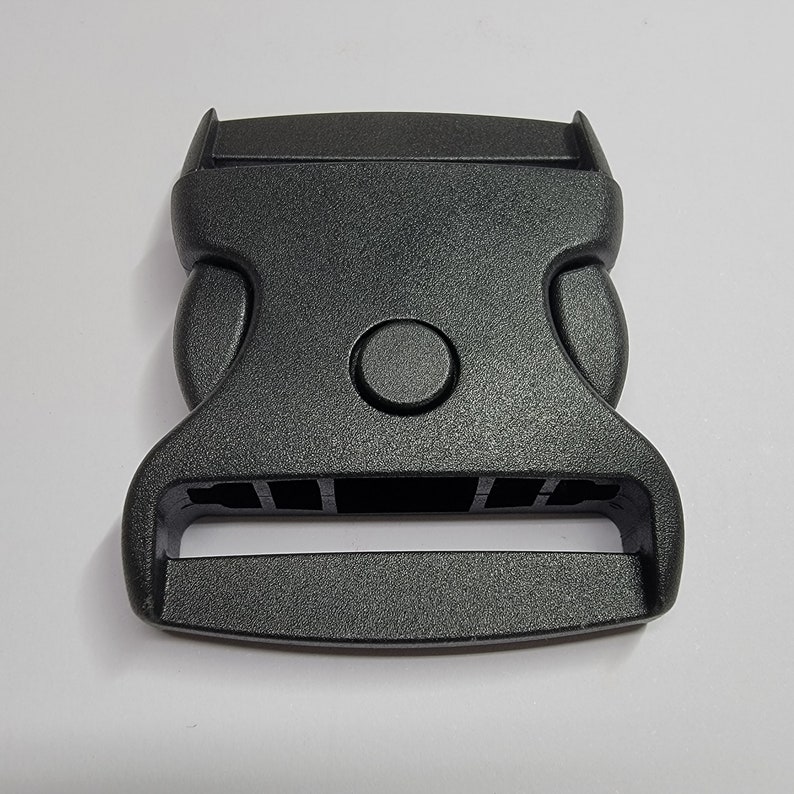 May include: A black plastic buckle with a smooth, textured surface. The buckle has a rectangular opening for a strap and a round button on the top.