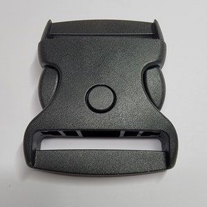 May include: Black plastic buckle with a smooth, matt finish. The buckle has a rectangular shape with rounded edges and a single, round button on the top.