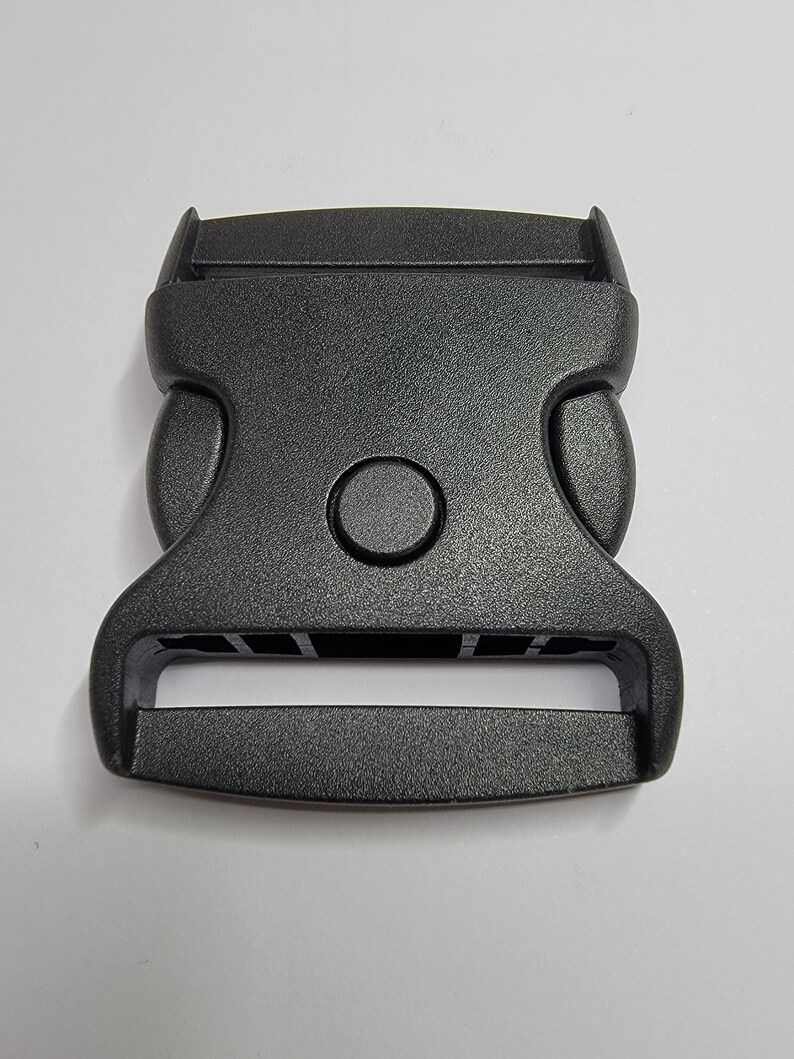 May include: Black plastic buckle with a smooth, matt finish. The buckle has a rectangular shape with rounded edges and a single, round button on the top.
