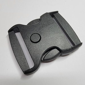 May include: A black plastic buckle with a side release mechanism. The buckle is designed for use with webbing or straps.