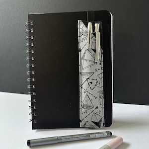 May include: A black spiral bound notebook with a grey and white geometric patterned pen holder attached to the cover. The pen holder has two pens inside. There are two pens lying on a white surface below the notebook.