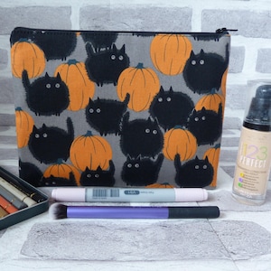 May include: A grey zippered pouch with a black and orange print of cats and pumpkins. The pouch is lying on a white surface with a set of coloured pencils, a makeup brush, and a bottle of foundation.