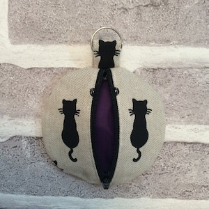 May include: A small, round, zippered pouch with a black cat print on a beige background. The pouch has a purple lining and a silver ring for attaching keys.
