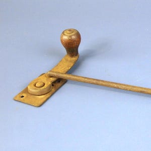 May include: An antique window operator with a wooden handle and a brass-colored metal frame. The operator has a long, slender arm extending from a rectangular base. The handle is a rounded shape, and the metal shows signs of age.