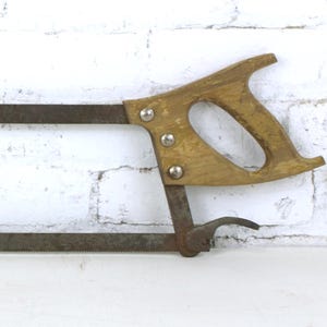 May include: A vintage hacksaw with a wooden handle and a metal frame. The handle is light brown and attached with three silver rivets. The blade is a dark, rusty color. The saw is designed for cutting metal and other materials.