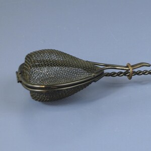 May include: A vintage tea infuser with a heart-shaped mesh basket, a twisted metal handle, and a wooden grip. The infuser is designed for steeping loose leaf tea. The handle is approximately 6 inches long.