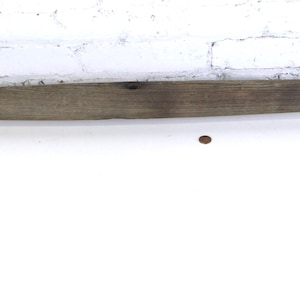 May include: A weathered wooden yoke with two metal rings. The yoke is a long, rectangular piece of wood with a slightly curved shape. The metal rings are attached to the ends of the yoke. The background is a white brick wall.