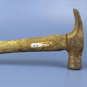 May include: A claw hammer with a weathered, light brown wooden handle and a rusted metal head. A small, partially worn white sticker with illegible black text is attached to the side of the wooden handle near the metal head of the tool.