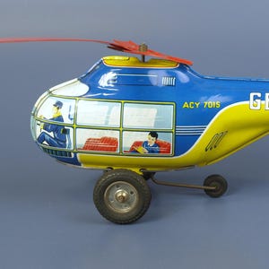 May include: Vintage toy helicopter in blue and yellow with a red propeller. The toy has a detailed cockpit with painted figures and the text "ACY 7015" and "GE 273". The helicopter is mounted on a single wheel and a small tail wheel.