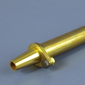 Peut inclure: Un outil cylindrique en laiton doté d'une buse conique à une extrémité, d'un petit boulon métallique fixant une bride circulaire près de la pointe et d'un bouton de réglage moleté à l'autre extrémité. La surface métallique a une finition dorée.