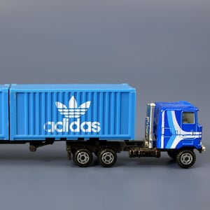 Puede incluir: Un camión de juguete semi-remolque azul con dos contenedores azules. Los contenedores tienen el logotipo de Adidas y la palabra "adidas" impresa en ellos.