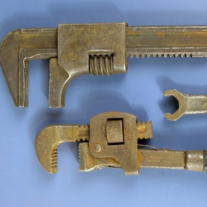 May include: Three vintage wrenches, including a pipe wrench with a black handle, a wrench with a serrated jaw, and a wrench with a smooth jaw. The wrenches are all made of metal and have a rusty patina.