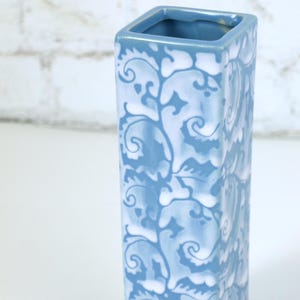 Op de afbeelding: Een hoge, rechthoekige keramische vaas met een lichtblauw en wit bloemmotief. De vaas heeft een vierkante opening en is ongeveer 20 cm hoog. Het ontwerp toont wervelende, gestileerde bladeren en ranken.