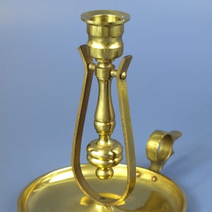 Op de afbeelding: Een vintage messing kandelaar met een ronde basis, een centrale stam en een gebogen handvat. De kandelaar heeft een klassiek ontwerp met een komvormige houder voor de kaars. Het messing heeft een gepolijste afwerking.