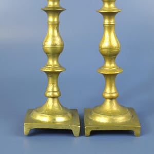 May include: Two antique brass candlesticks. Each candlestick has a square base, a slender, ornate stem, and a flared top. The brass has a warm, aged patina, and the candlesticks are displayed against a blue background.