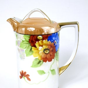 Puede incluir: Una jarra de cerámica blanca con un borde dorado y un diseño floral. La jarra tiene una tapa con un asa dorada. Las flores son rojas, amarillas, azules y verdes.