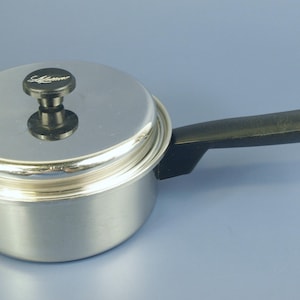 Puede incluir: Una pequeña cacerola de acero inoxidable con tapa y asa negra. La tapa tiene un pomo negro con la palabra "Saladmaster". El asa tiene un pequeño lazo de metal en el extremo. La cacerola está sobre una superficie azul claro.