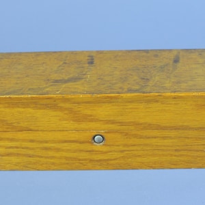 May include: A rectangular wooden box with a light brown finish. The box has a small, round metal clasp on the front. The wood grain is visible, and the box appears to be well-constructed. The box is likely used for storage.