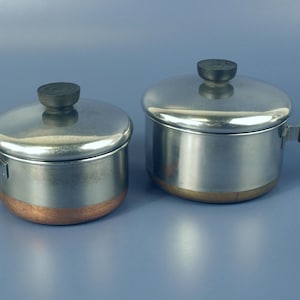Puede incluir: Dos cacerolas de acero inoxidable con bases de color cobre y tapas a juego. Cada olla cuenta con un mango negro con una pequeña anilla metálica para colgar en el extremo y un pomo negro en la tapa. Las ollas están colocadas una al lado de otra.