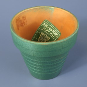 Puede incluir: Una maceta de arcilla verde texturizada con un interior naranja claro. Dentro de la maceta hay una etiqueta verde con texto blanco: NEW - IMPROVED - DRIPLESS HEALTH-O-PLANT CLAY POT WITH WATERING RESERVOIR MADE IN U.S.A. El exterior presenta estrías.