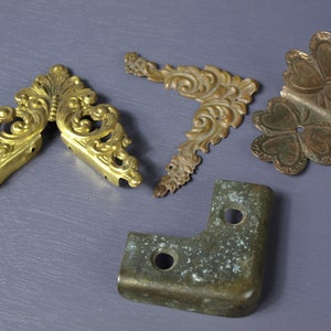 Puede incluir: Cuatro soportes de esquina de metal adornados en oro y bronce. Dos de los soportes están diseñados intrincadamente con patrones florales, mientras que los otros dos son más simples, uno con un diseño de trébol y el otro con una forma redonda simple.