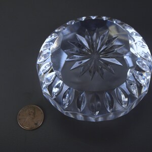 May include: A clear, round crystal ashtray with a starburst design in the center and a decorative leaf pattern around the edge. A penny is next to the ashtray for size comparison.