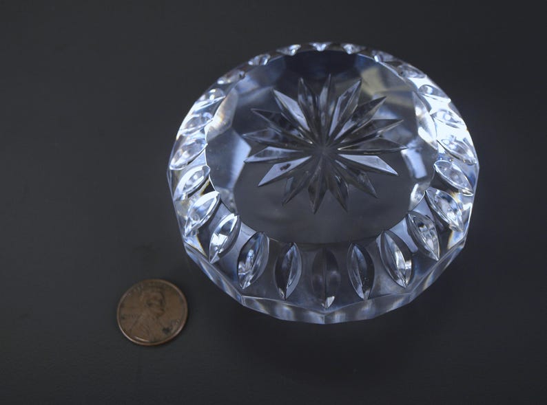 May include: A clear, round crystal ashtray with a starburst design in the center and a decorative leaf pattern around the edge. A penny is next to the ashtray for size comparison.