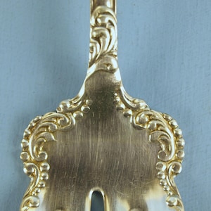 May include: Close-up of the ornate, gold-toned handle of a fork. The handle is decorated with a swirling, floral design.